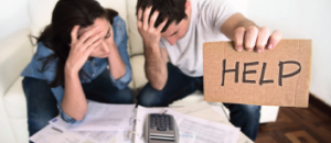 couple worried with their heads down as they look at their bills spread on a table with a calculator on top the husband holds up a cardboard sign written HELP