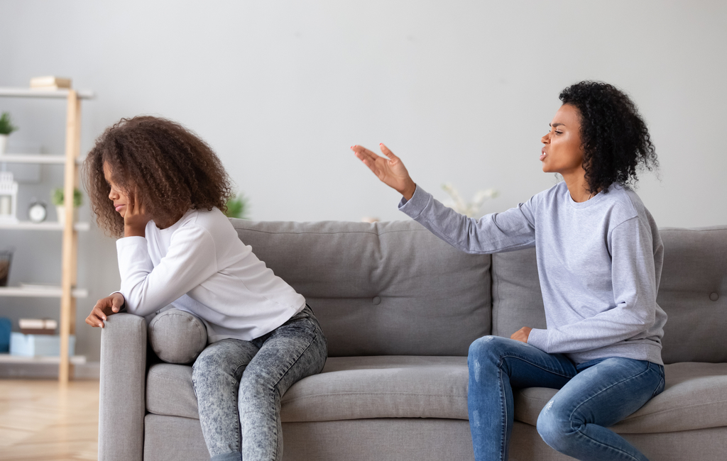 Mother arguing with teenager as teenage daughter has her back turned to her mom, both are sitting on the sofa